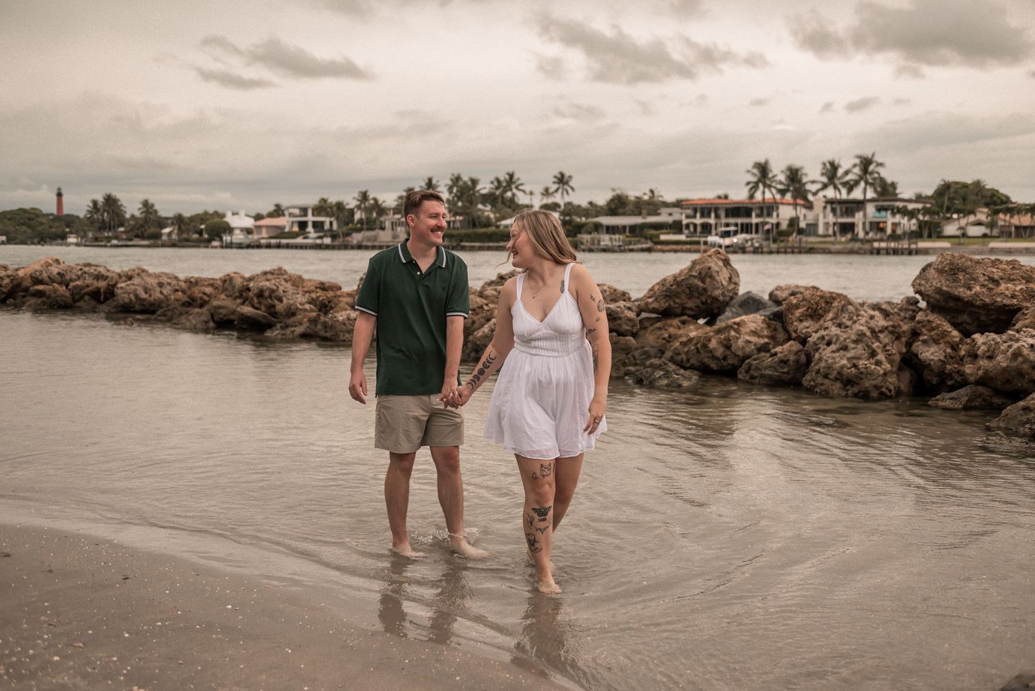 Couple walking hand in hand along a tropical beach at sunset with palm trees and boats in the background.