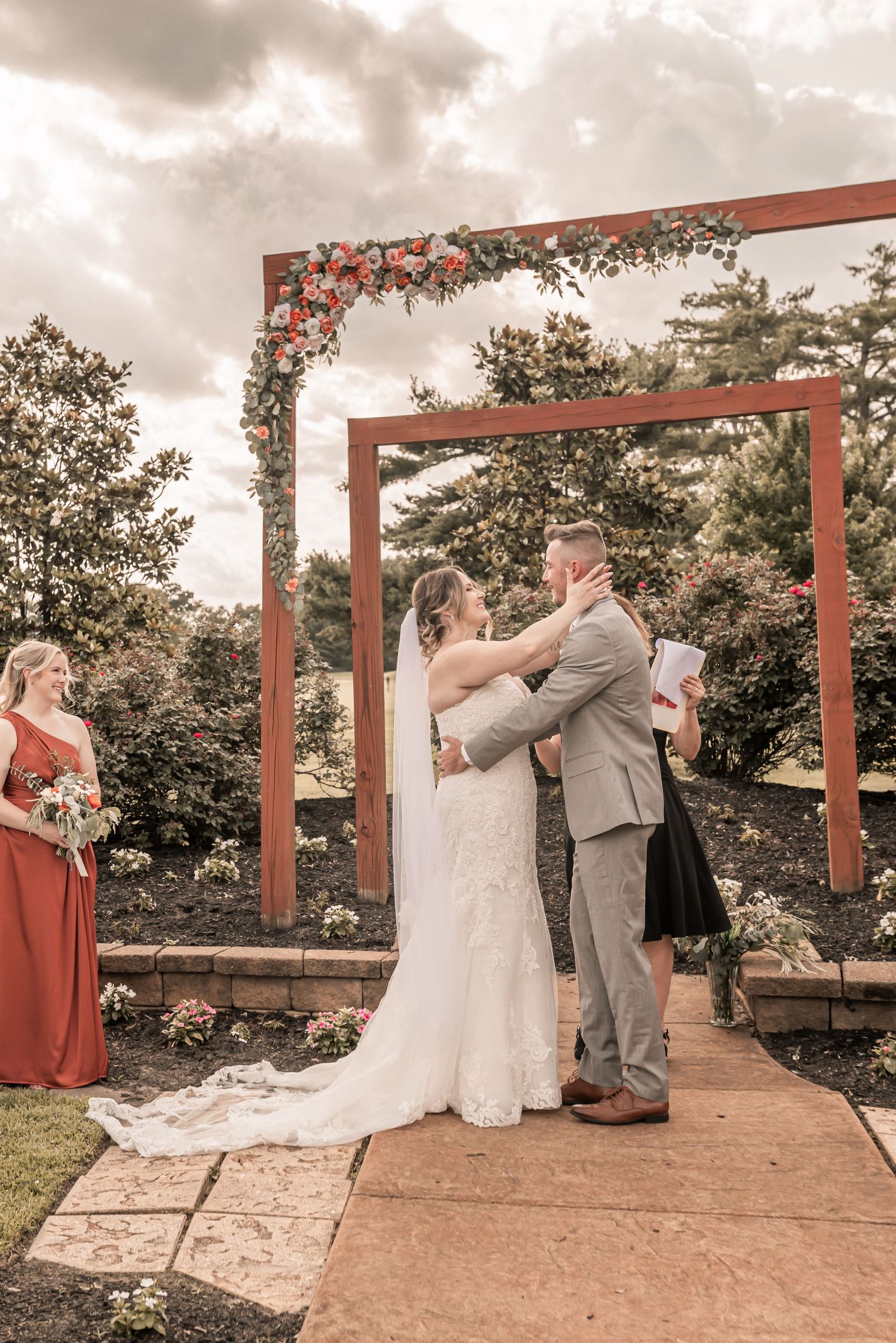 A wedding ceremony under a rustic wooden arch decorated with coral flowers against a moody cloudy sky.