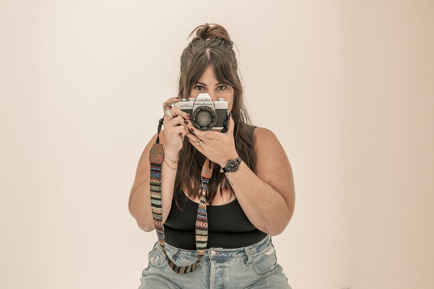 Mirror selfie showing someone in a black tank top and denim overalls holding a camera in a beige-toned room.