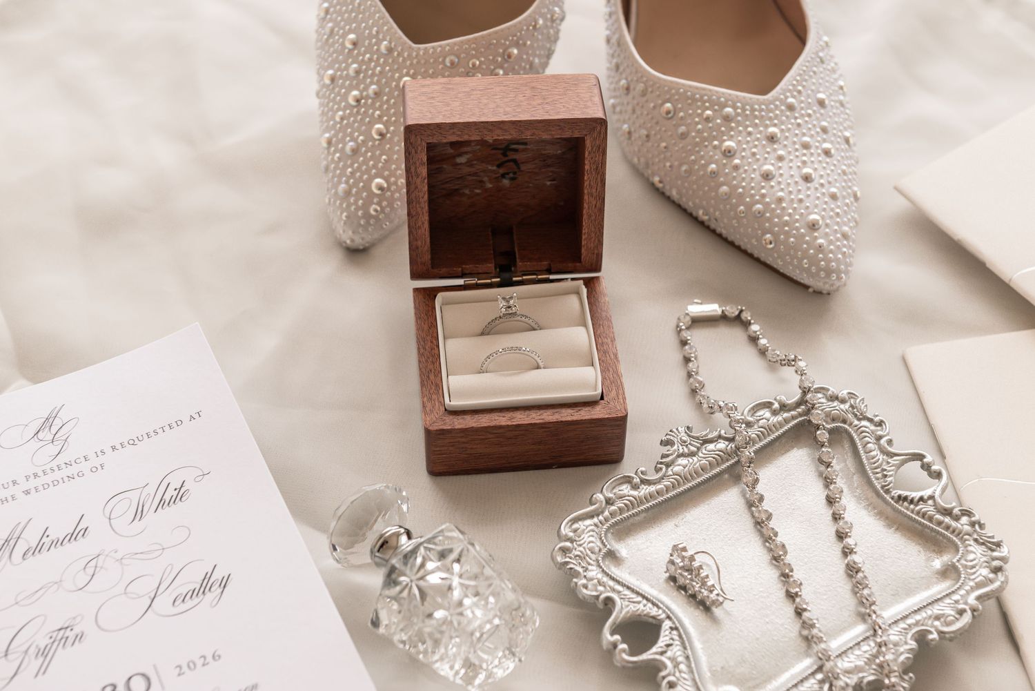 Elegant wedding flat lay featuring pearl bridal shoes, diamond ring in wooden box, crystal perfume bottle, and silver jewelry tray.
