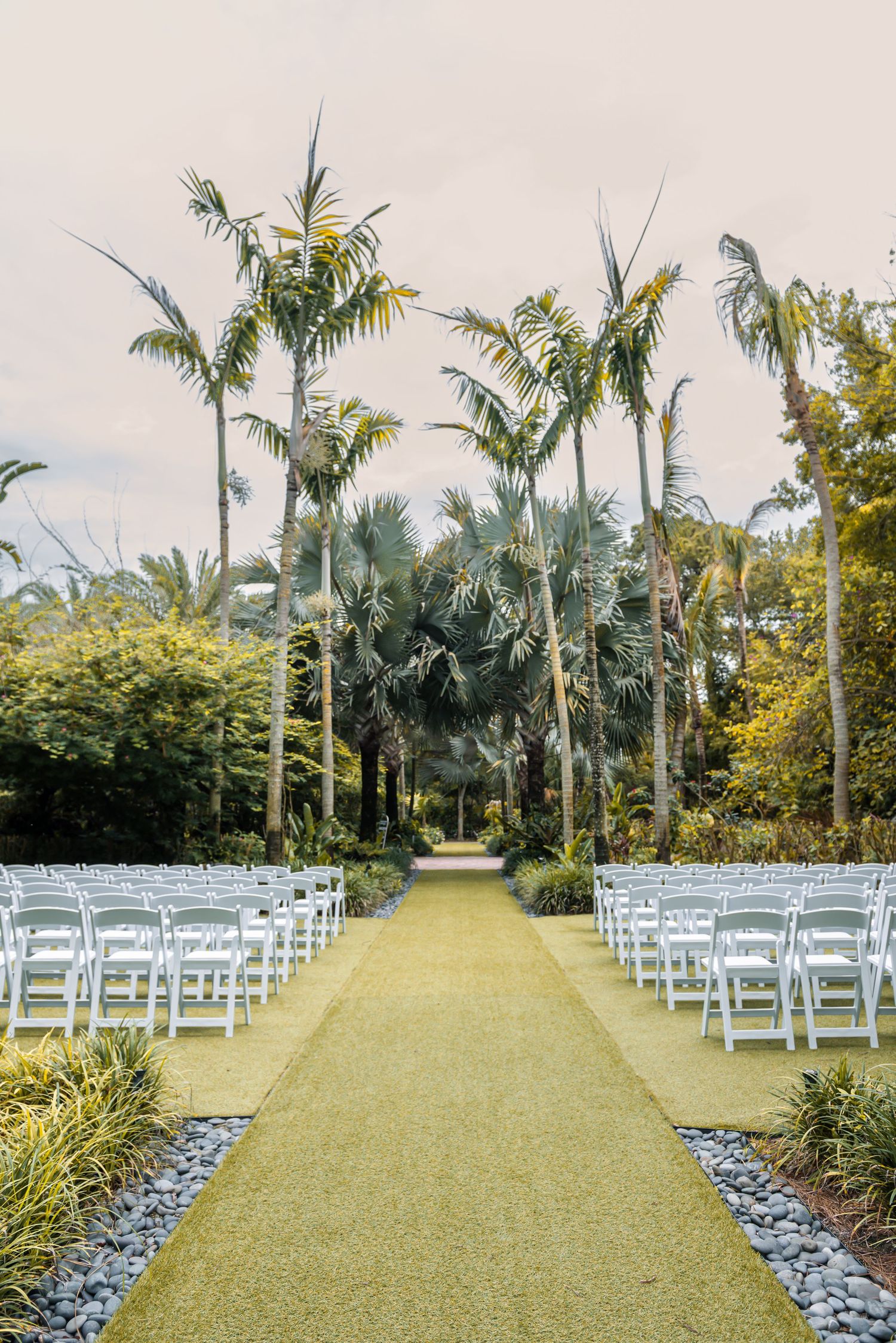 Outdoor wedding ceremony setup with white folding chairs lining a grass aisle surrounded by tall palm trees.
