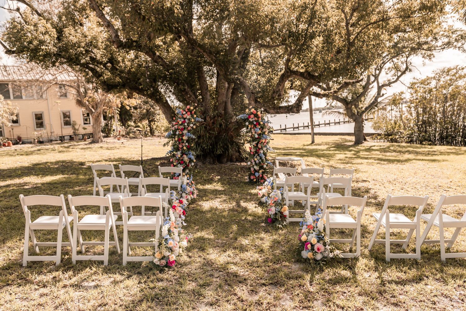 Outdoor wedding ceremony setup with white chairs and floral arch under a large oak tree.