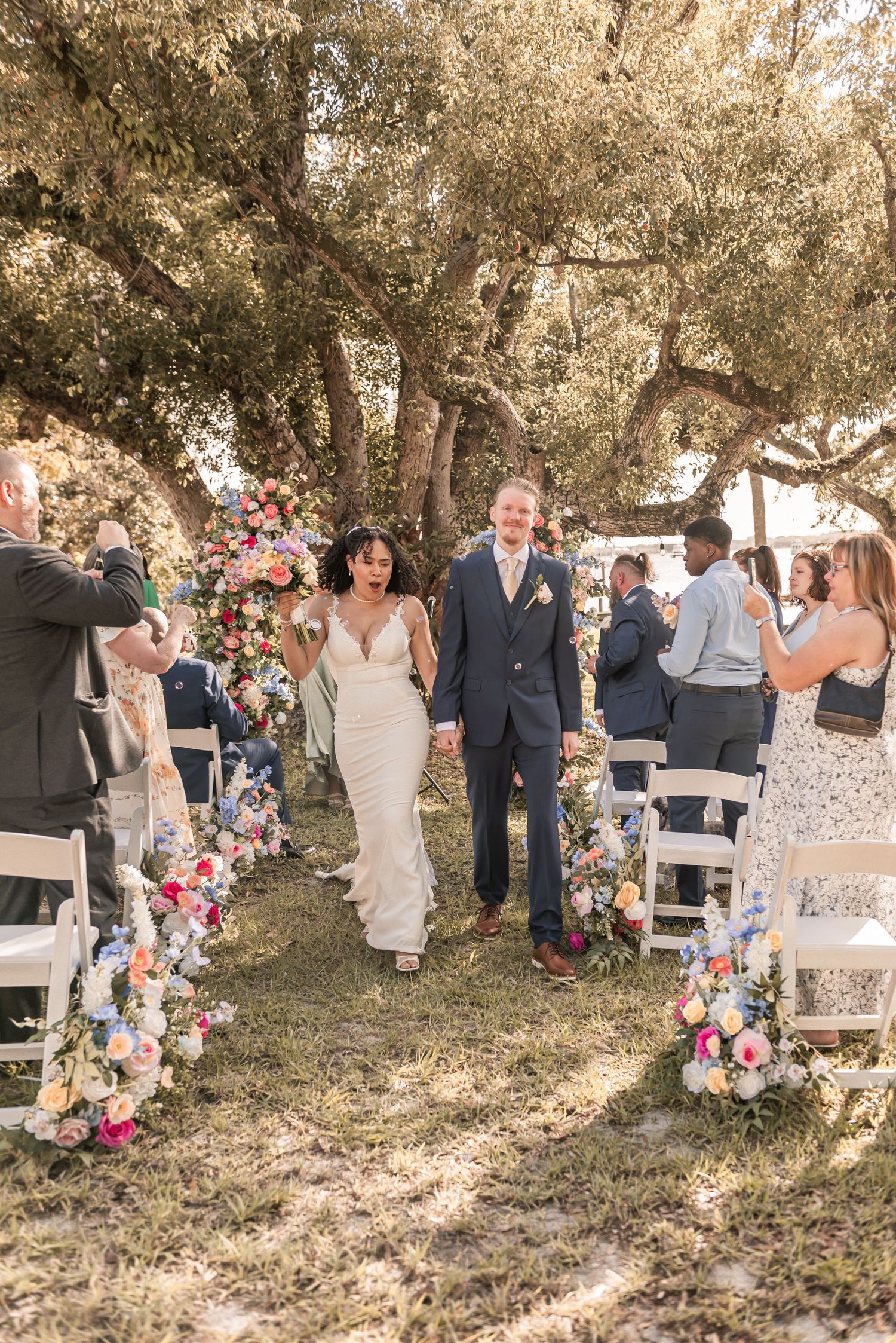 Newlyweds walk down floral-lined outdoor aisle under oak trees after wedding ceremony.