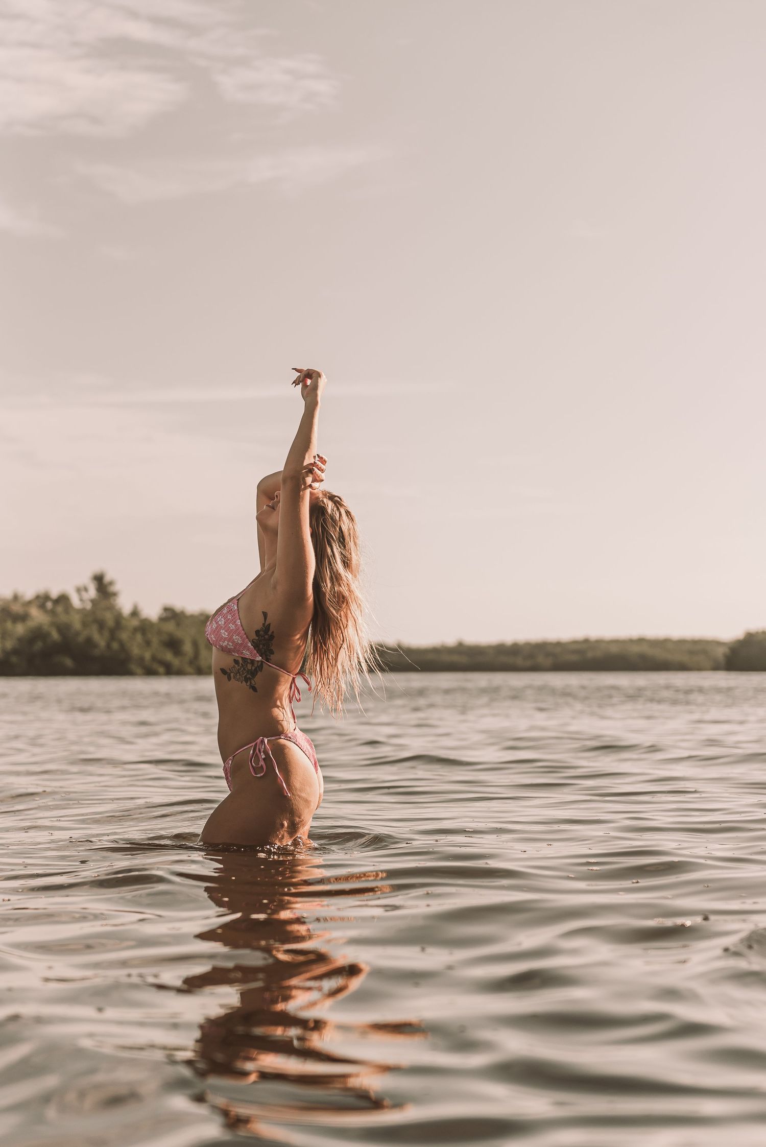Woman in pink bikini standing waist-deep in calm lake water, arm raised, long hair flowing, trees in background.