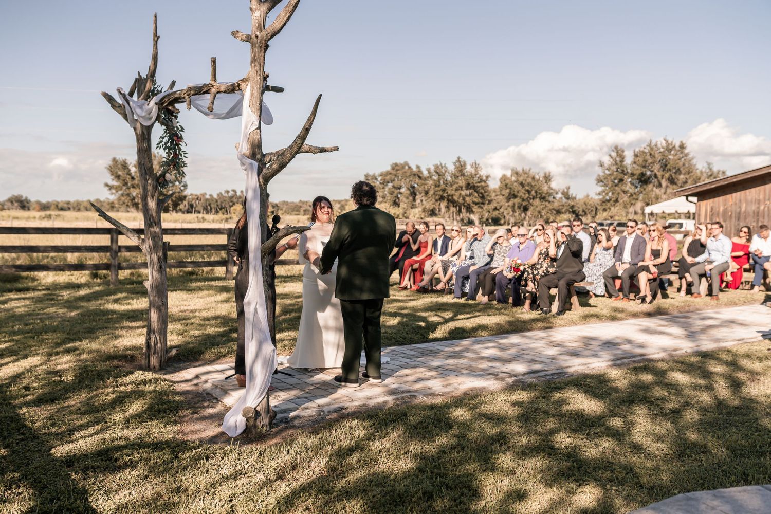 Panoramic view of an outdoor wedding ceremony with guests seated on wooden benches under blue skies.