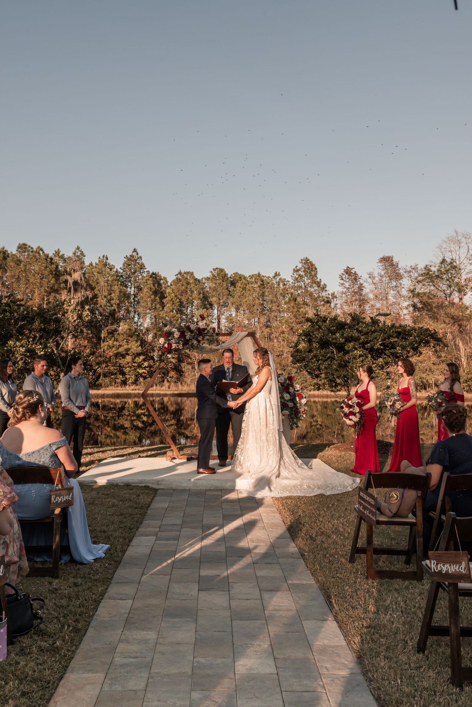An outdoor wedding ceremony takes place on a stone path with guests seated on wooden chairs at sunset.