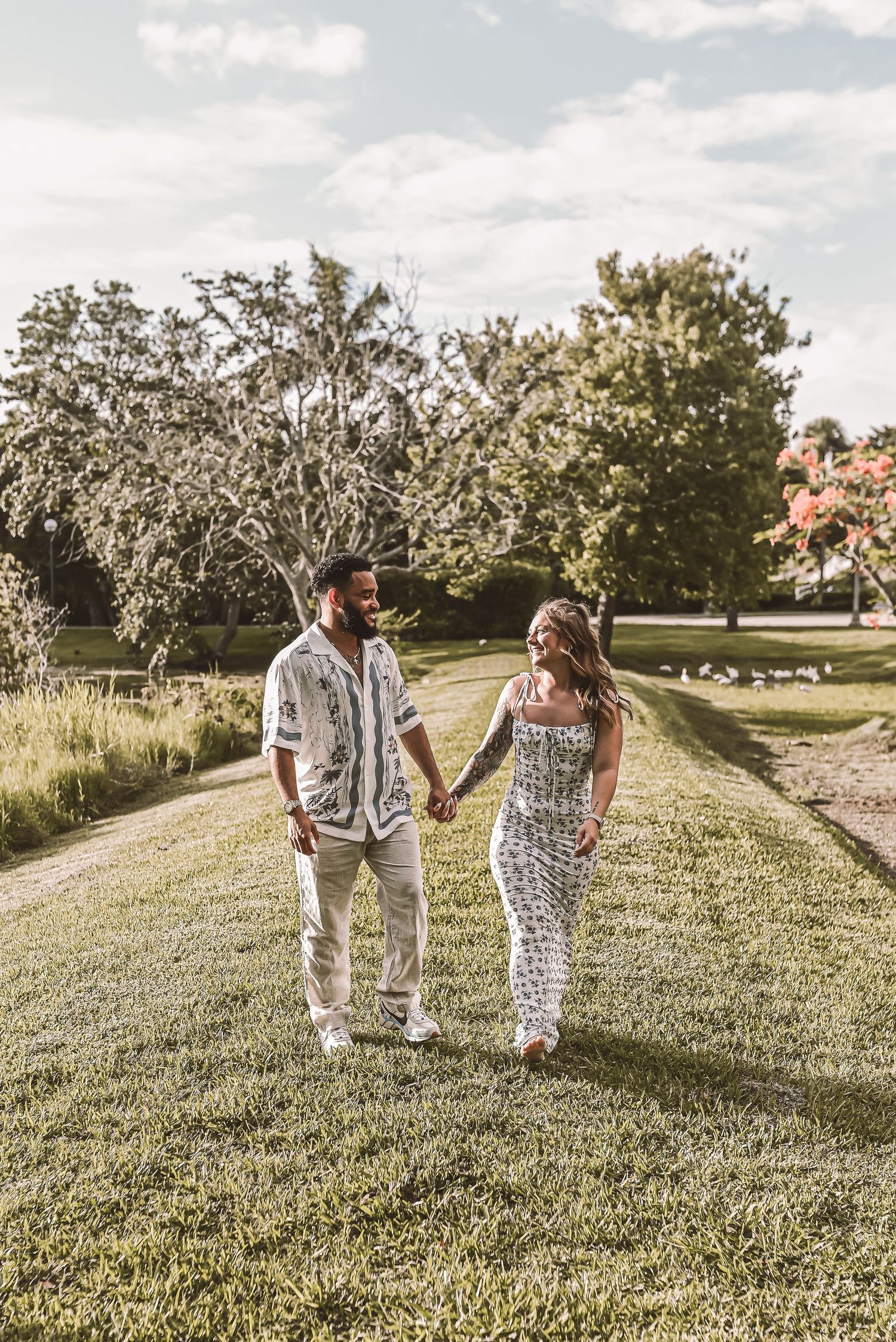 A couple walking hand in hand on a grassy path during golden hour with trees and sunlight in the background.