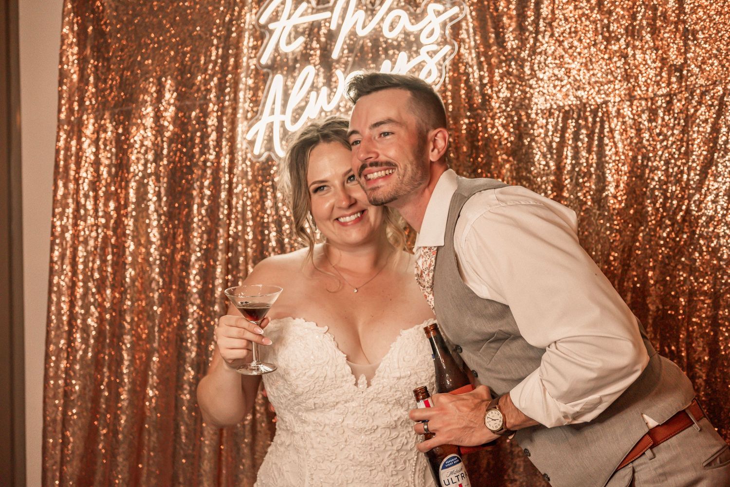 Two people pose playfully in front of a rose gold sequin backdrop at a festive celebration.