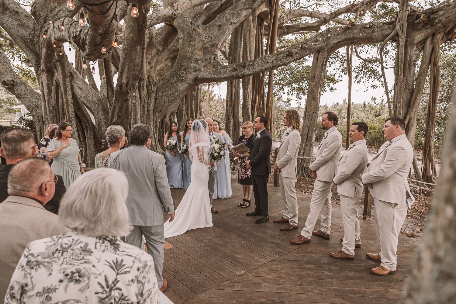 A panoramic sequence of an outdoor wedding ceremony under banyan trees with guests dressed in white attire.