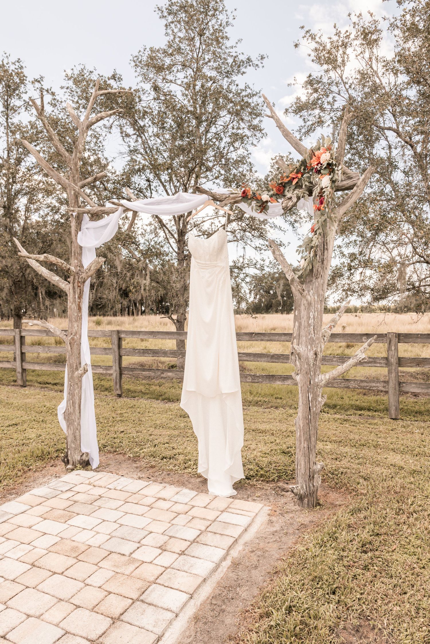 A white wedding dress hangs on a rustic wooden arch decorated with floral arrangements in an outdoor garden setting.