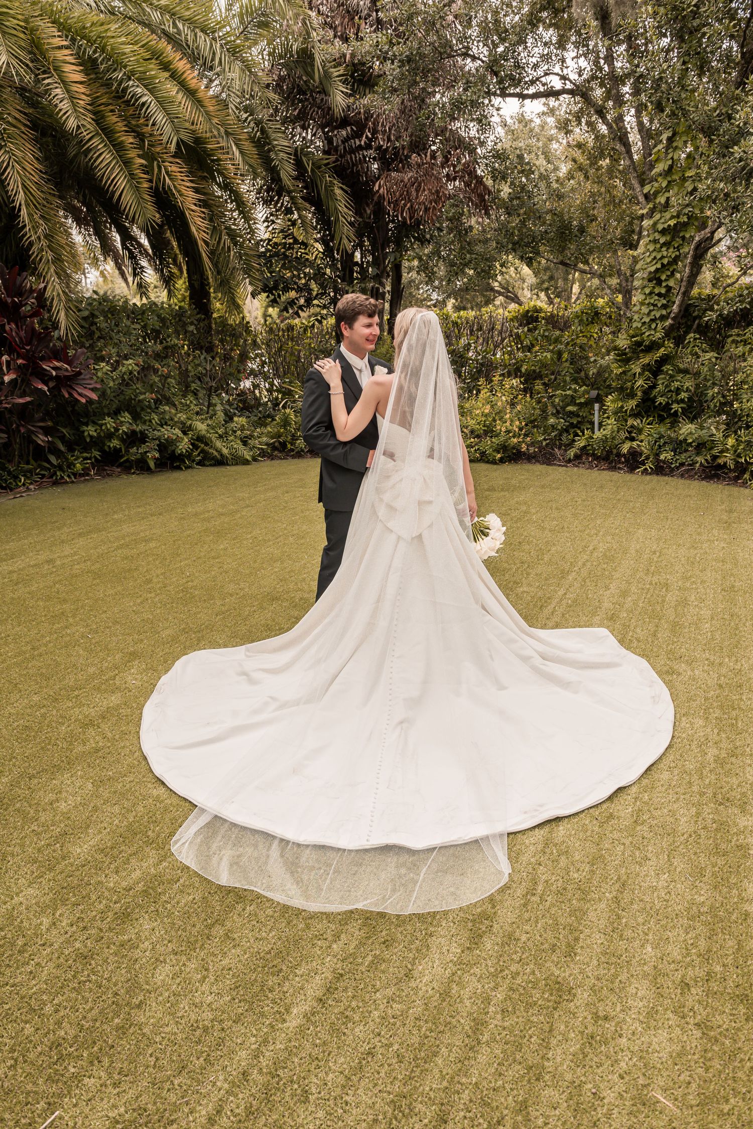 Bride in flowing white gown with cathedral veil dancing with groom in lush green garden setting.