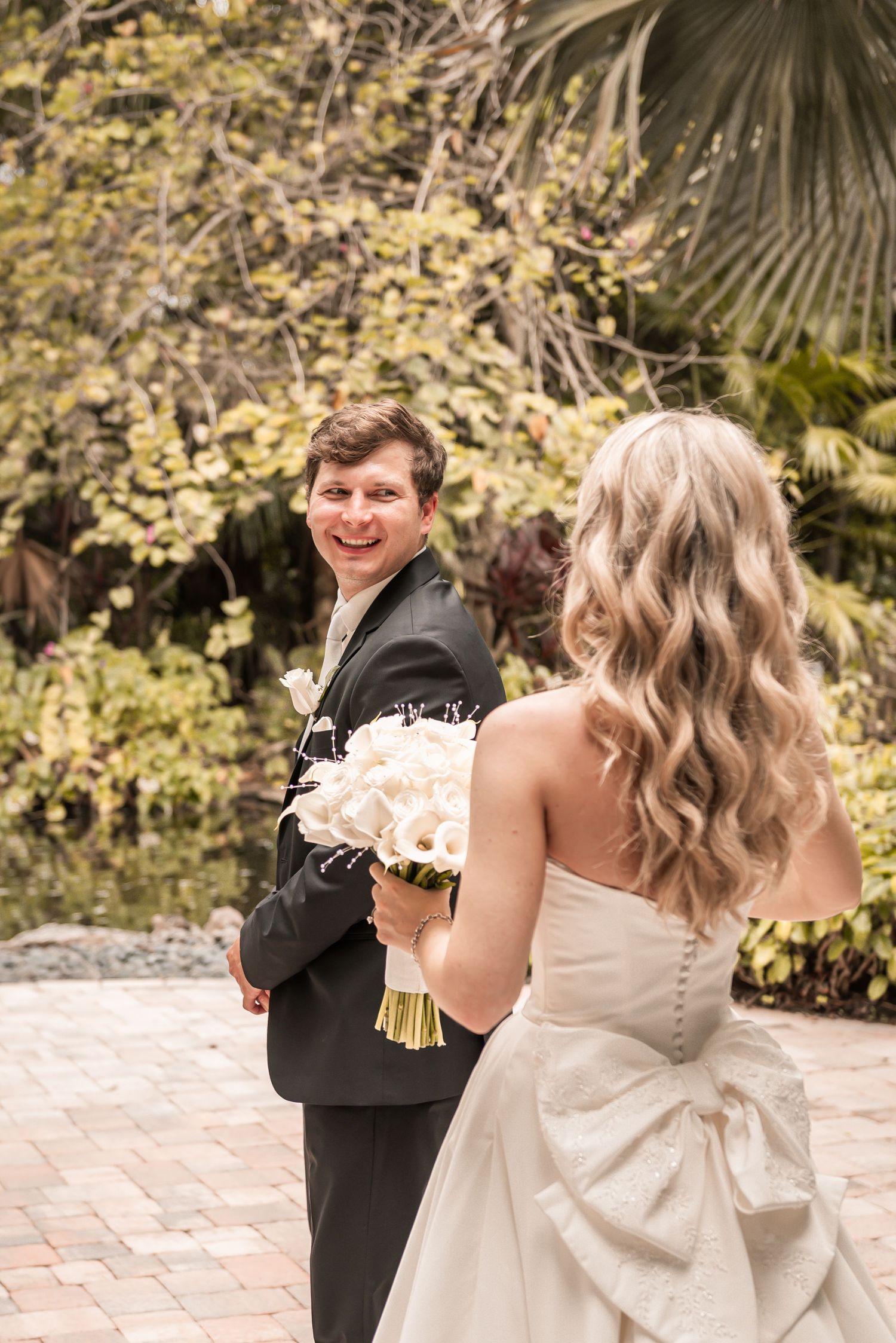 Groom smiling at bride in white strapless gown holding white bouquet during outdoor wedding first look.