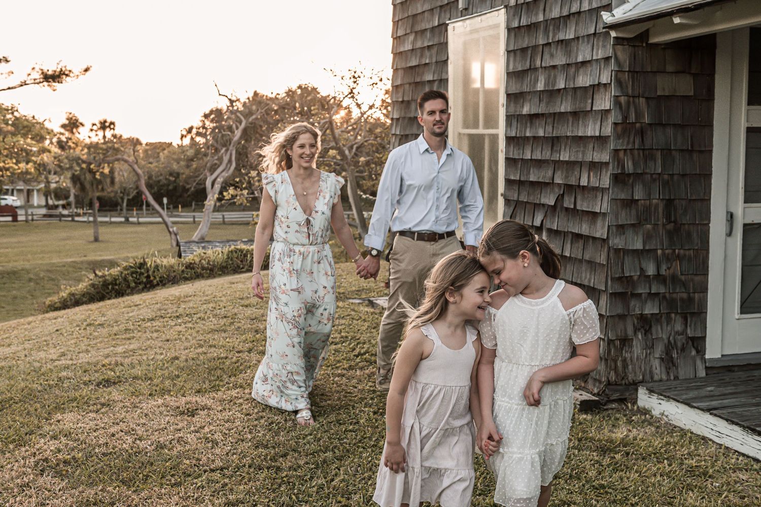 A couple in white outfits poses together in front of a rustic shingle-sided house during golden hour.