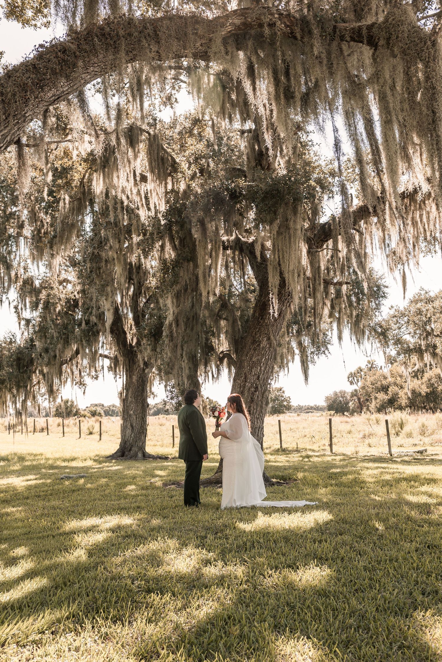 A wedding couple stands beneath Spanish moss-draped oak trees in a romantic Southern garden setting.