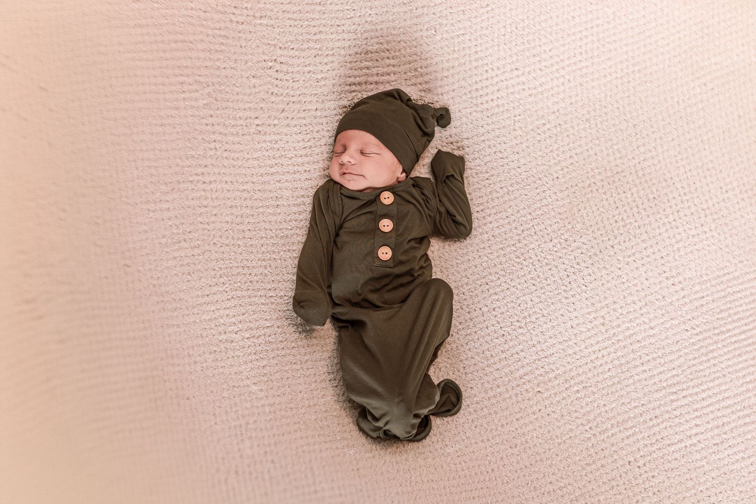 Newborn baby in olive green knit romper and knotted hat lying on cream textured blanket.