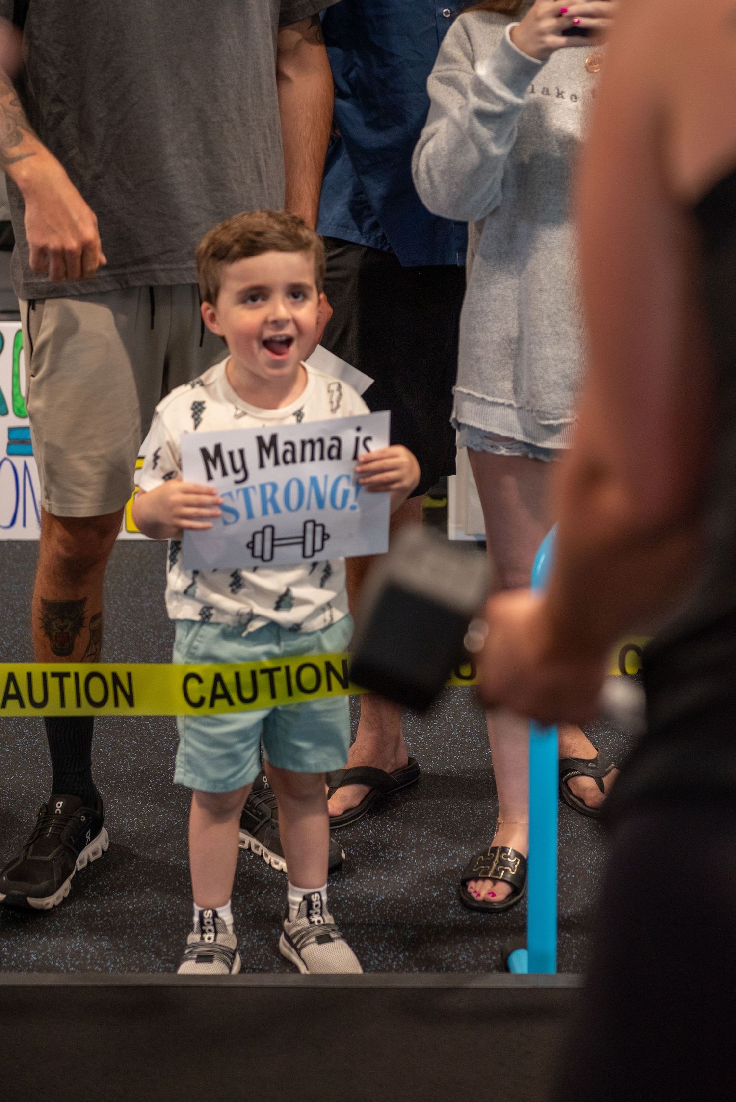 Excited young boy holds a 'My Mama is STRONG' sign at a weightlifting competition near caution tape.