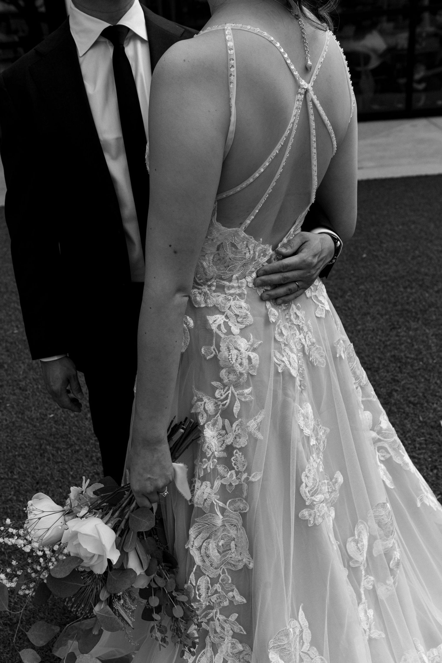 Black and white wedding photo showing intricate lace dress with delicate strappy back detail and floral bouquet.