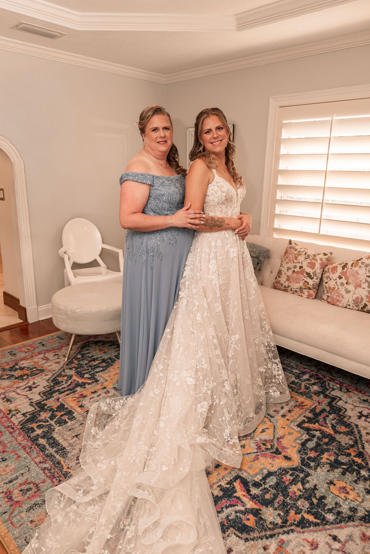 Two women in formal gowns, one in blue and one in white lace, stand together in a room with a patterned rug.