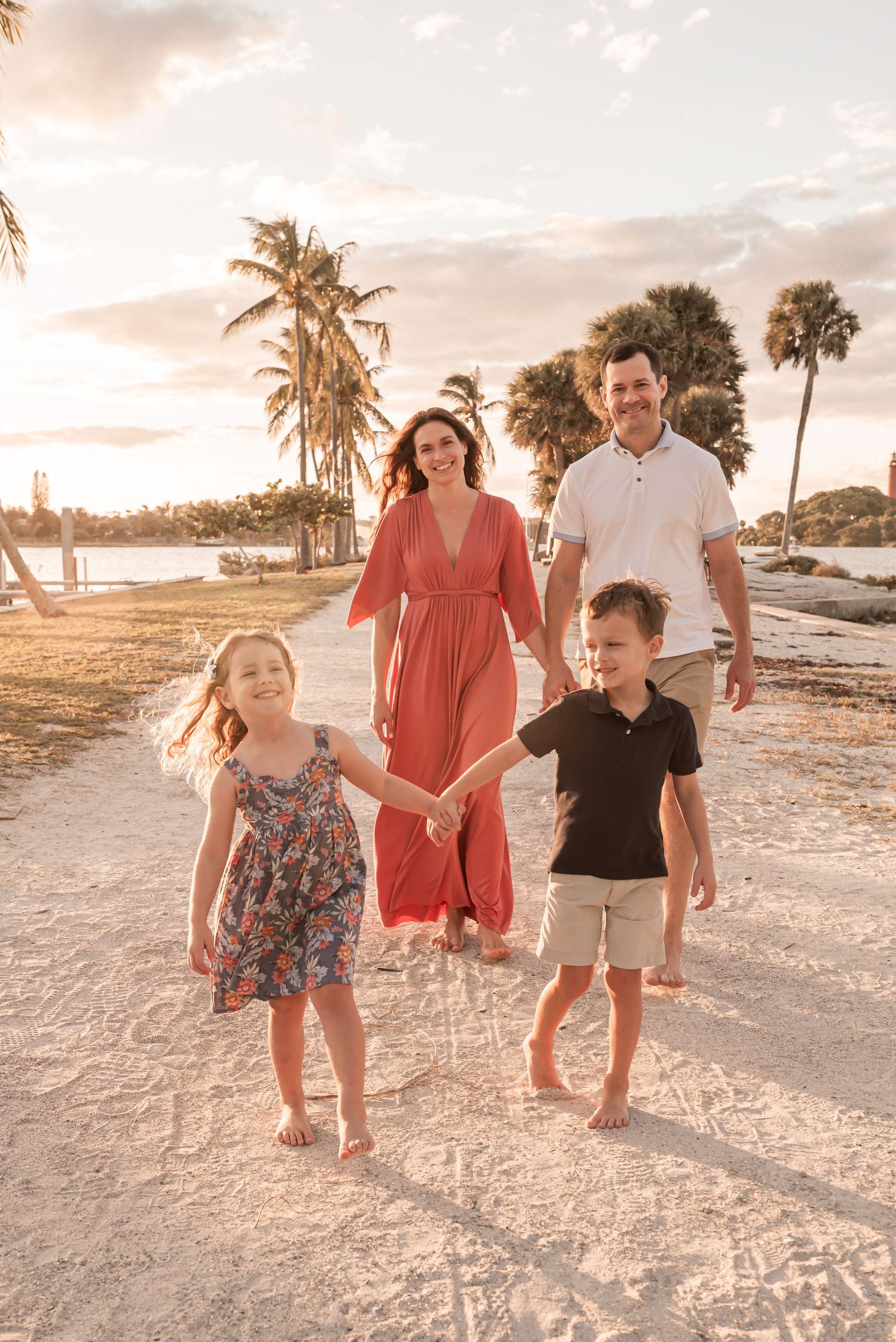 Family walking together on white sandy beach at sunset with palm trees and ocean in background.