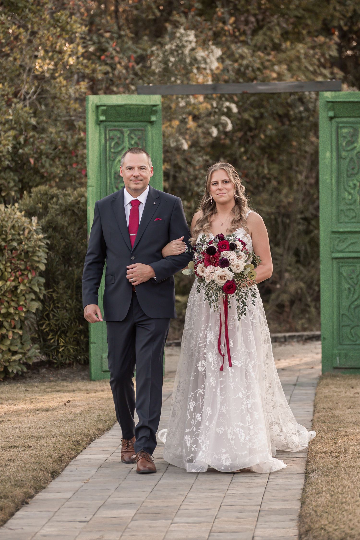 A wedding couple walks down a stone path with green doors in the background during an outdoor autumn ceremony.