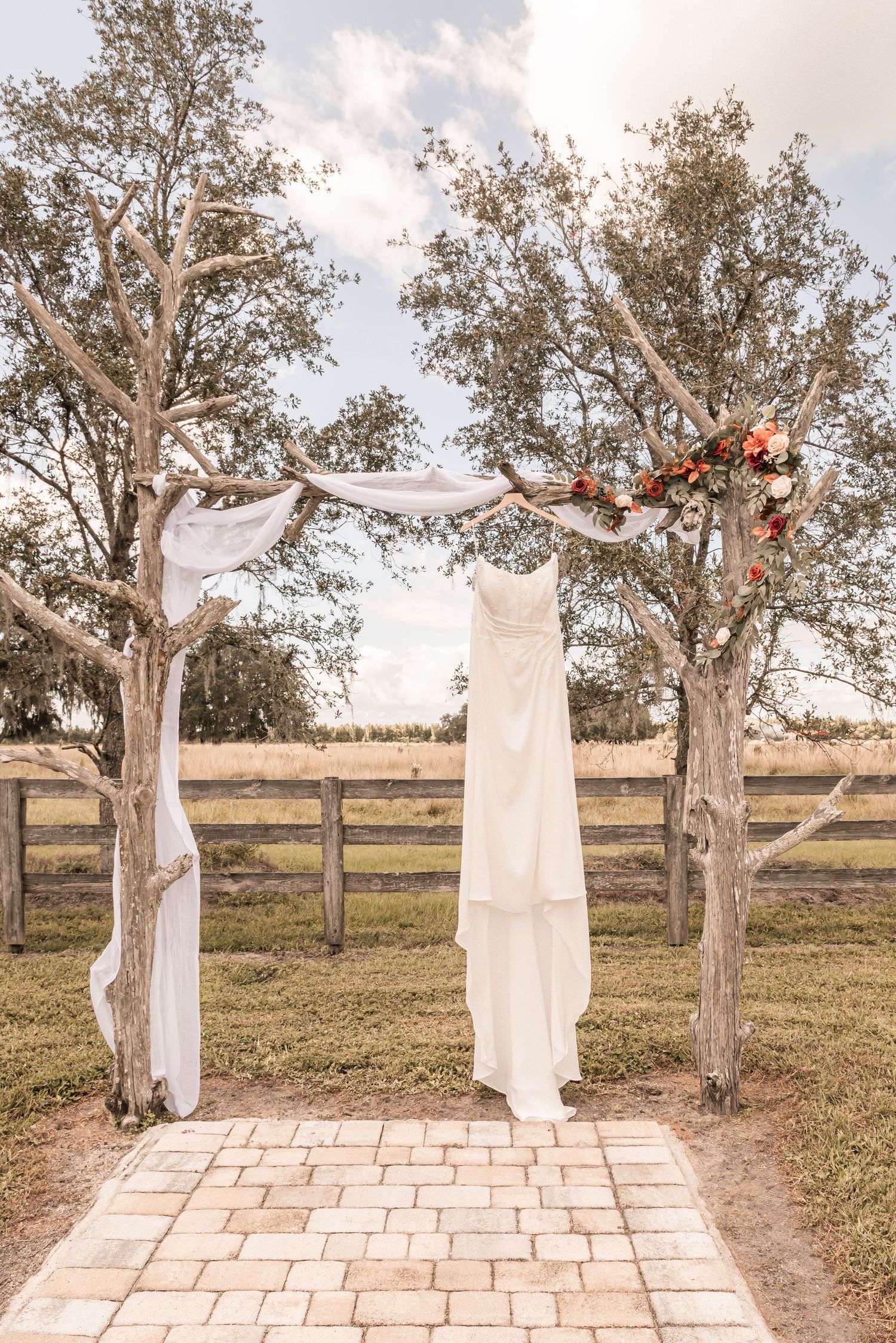 A white wedding dress hangs elegantly between two trees with draped fabric creating a rustic outdoor ceremony arch.