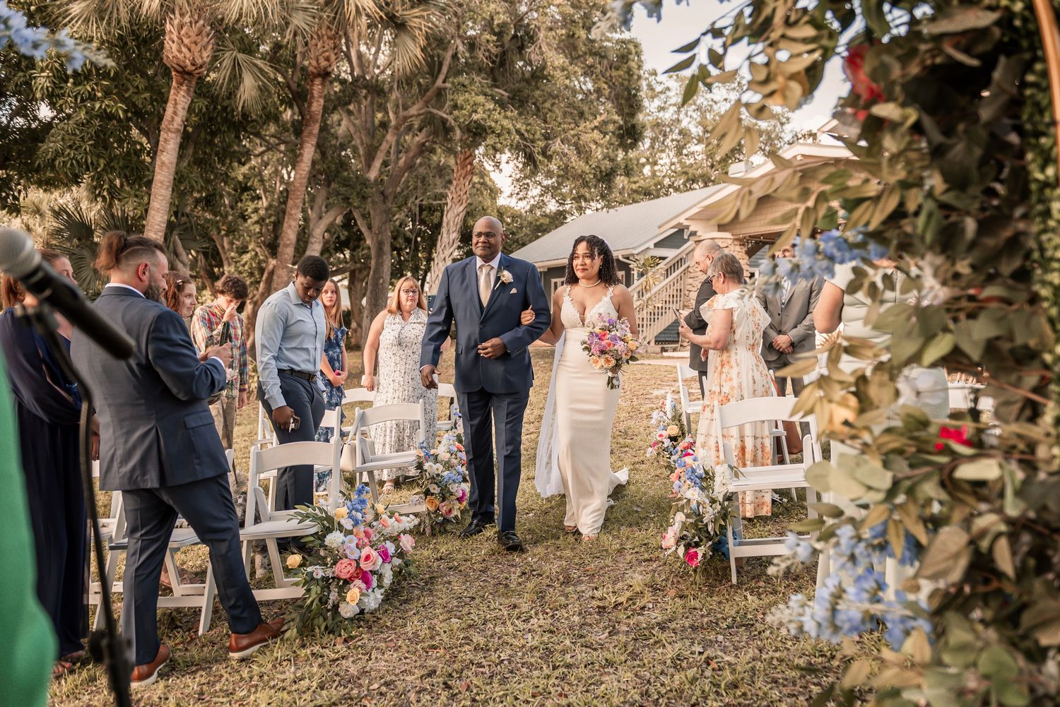 Newlywed couple walks down outdoor wedding aisle surrounded by guests, floral arrangements, and lush trees.
