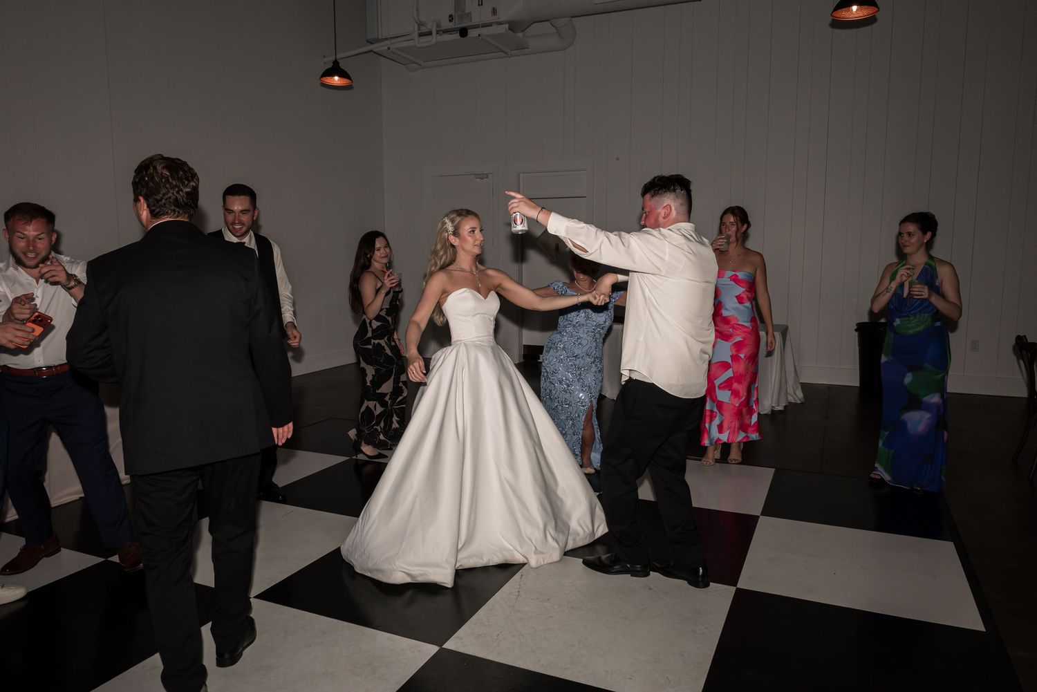 Bride and groom share first dance on checkered black and white floor surrounded by wedding guests.