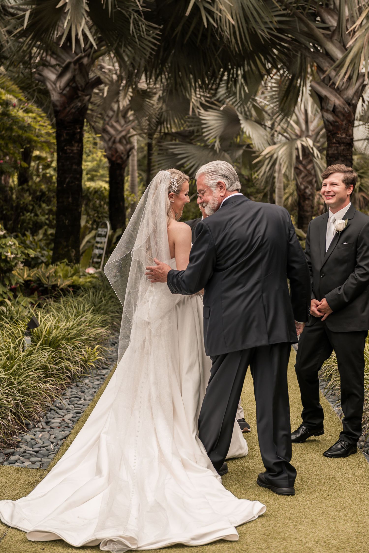 Bride in white gown and veil shares emotional moment with older man in dark suit at outdoor garden wedding ceremony.