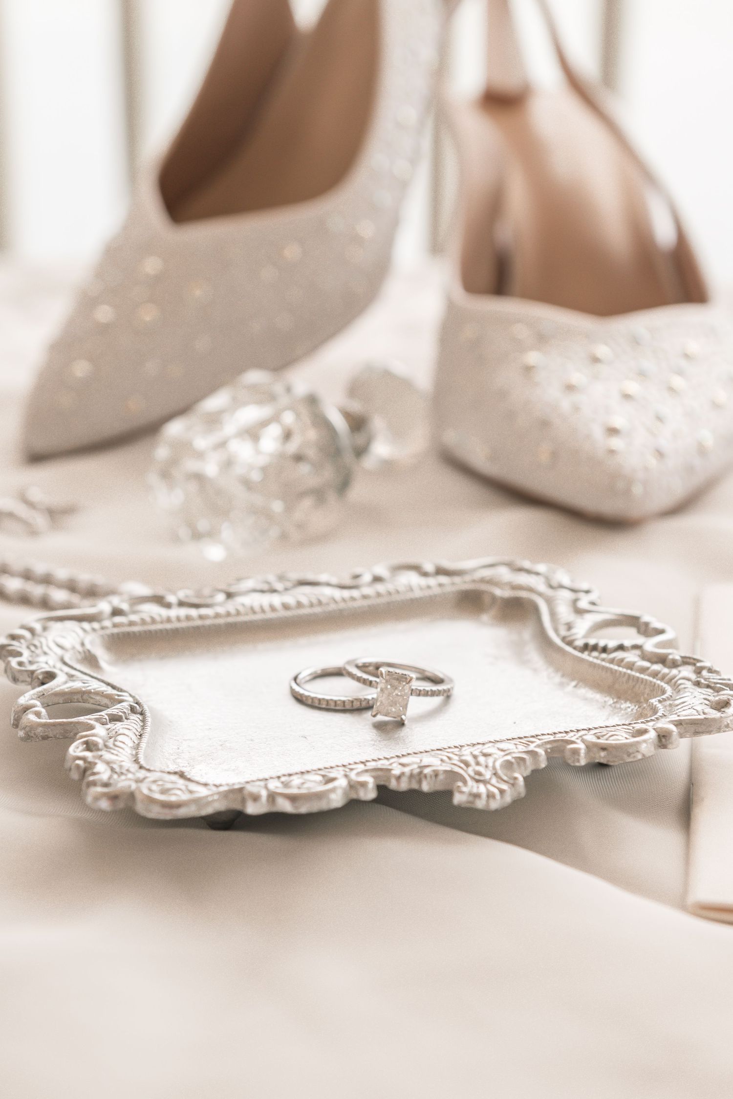 Elegant bridal flat lay with pearl-embellished white heels, diamond rings on ornate silver tray, and crystal perfume bottle.