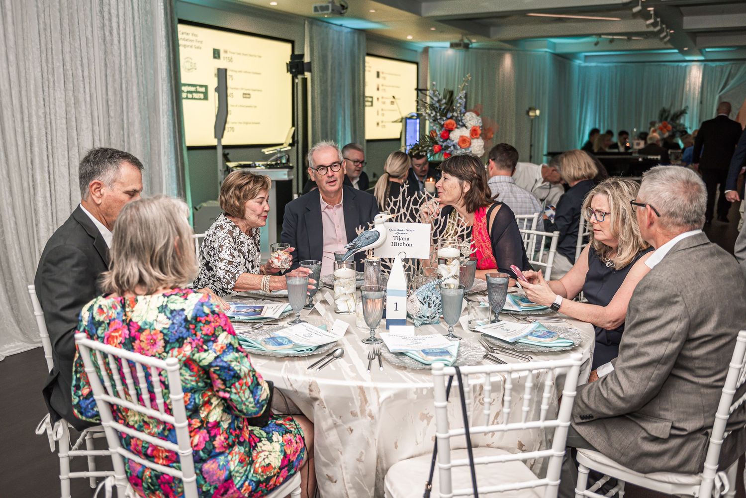 Guests seated at an elegantly decorated round table for a formal dining event with teal-colored walls in the background.