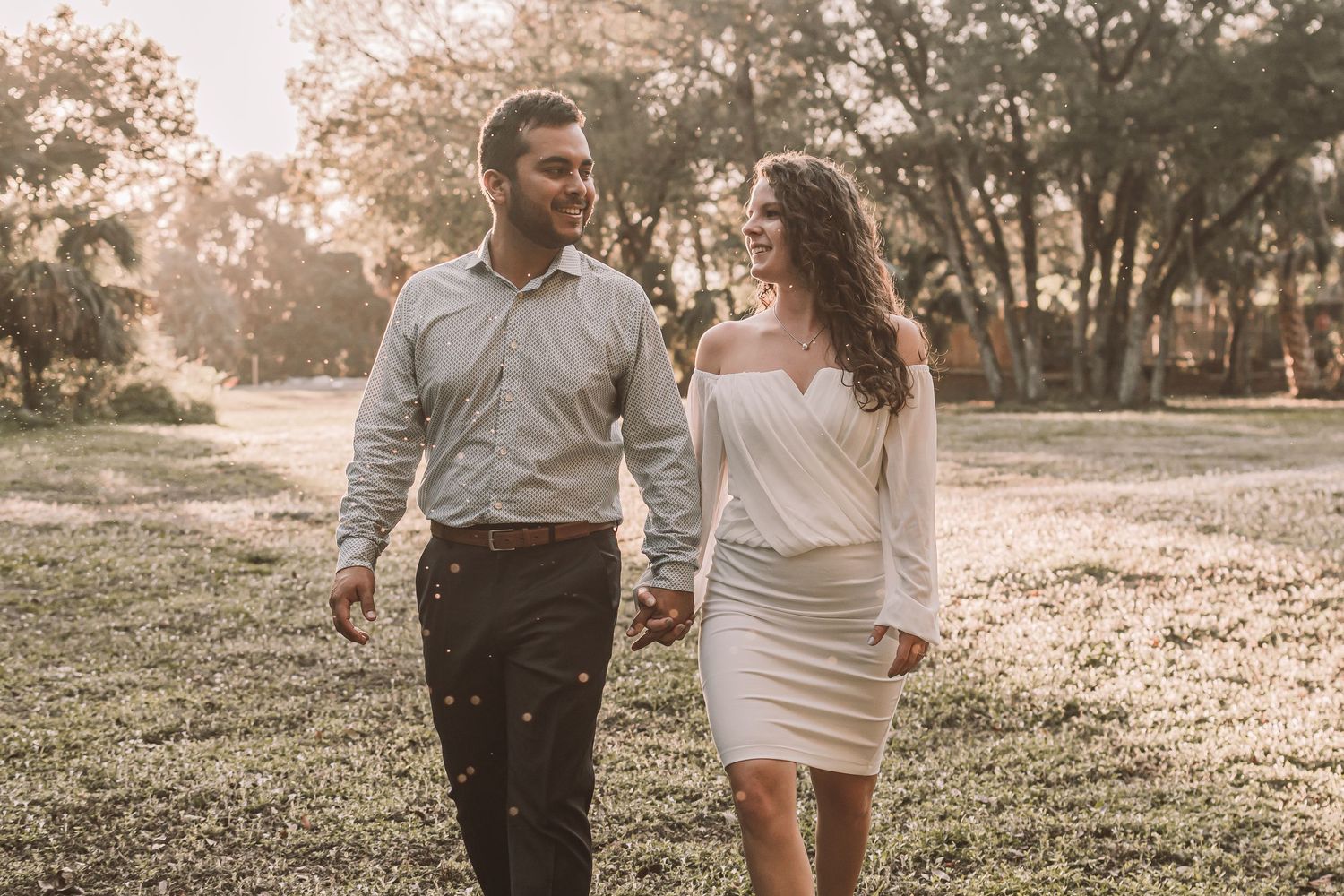 A couple holds hands while walking through a sun-dappled park during golden hour with trees in the background.