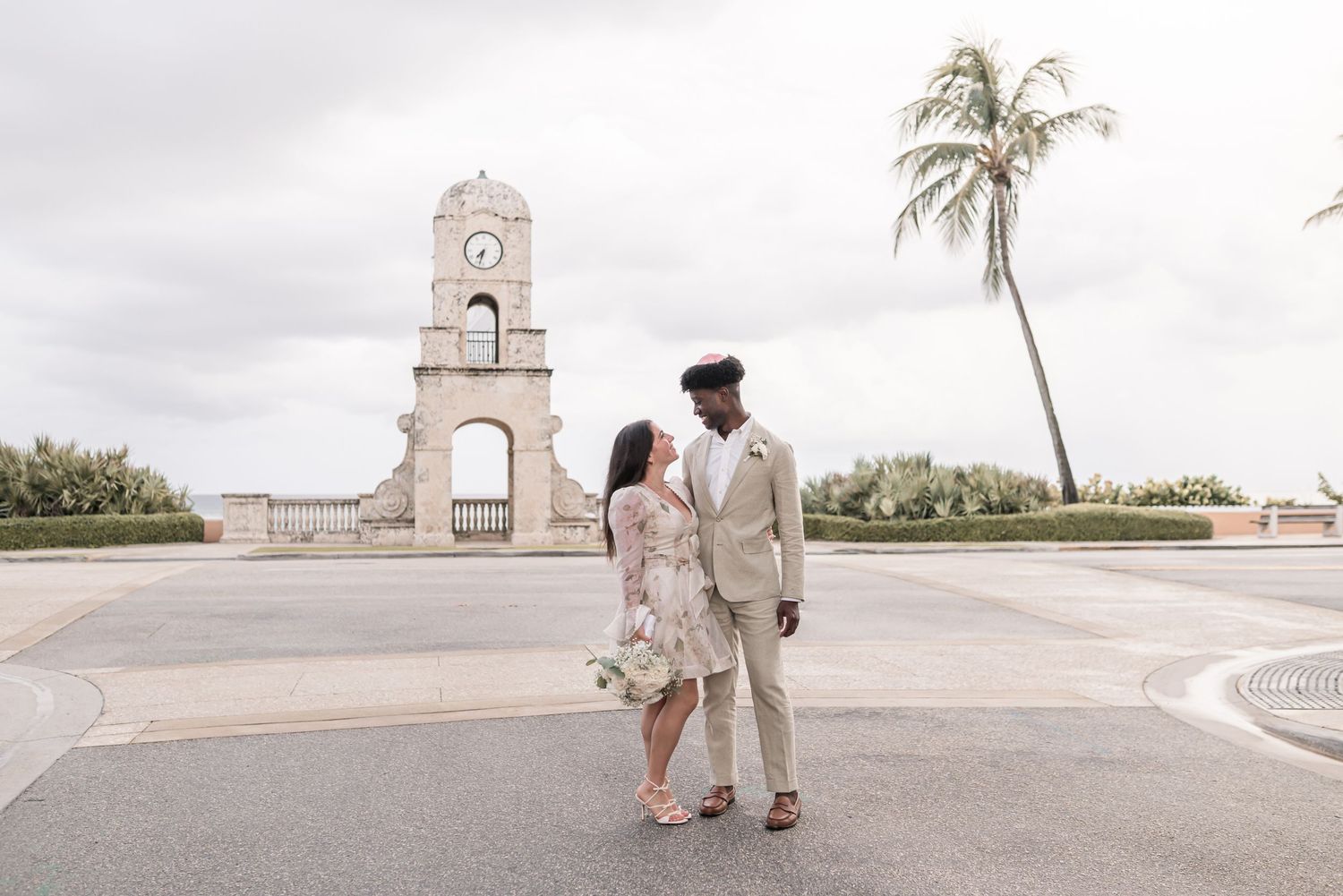 A couple stands in front of the historic Worth Avenue Clock Tower in Palm Beach on a cloudy day with a palm tree backdrop.