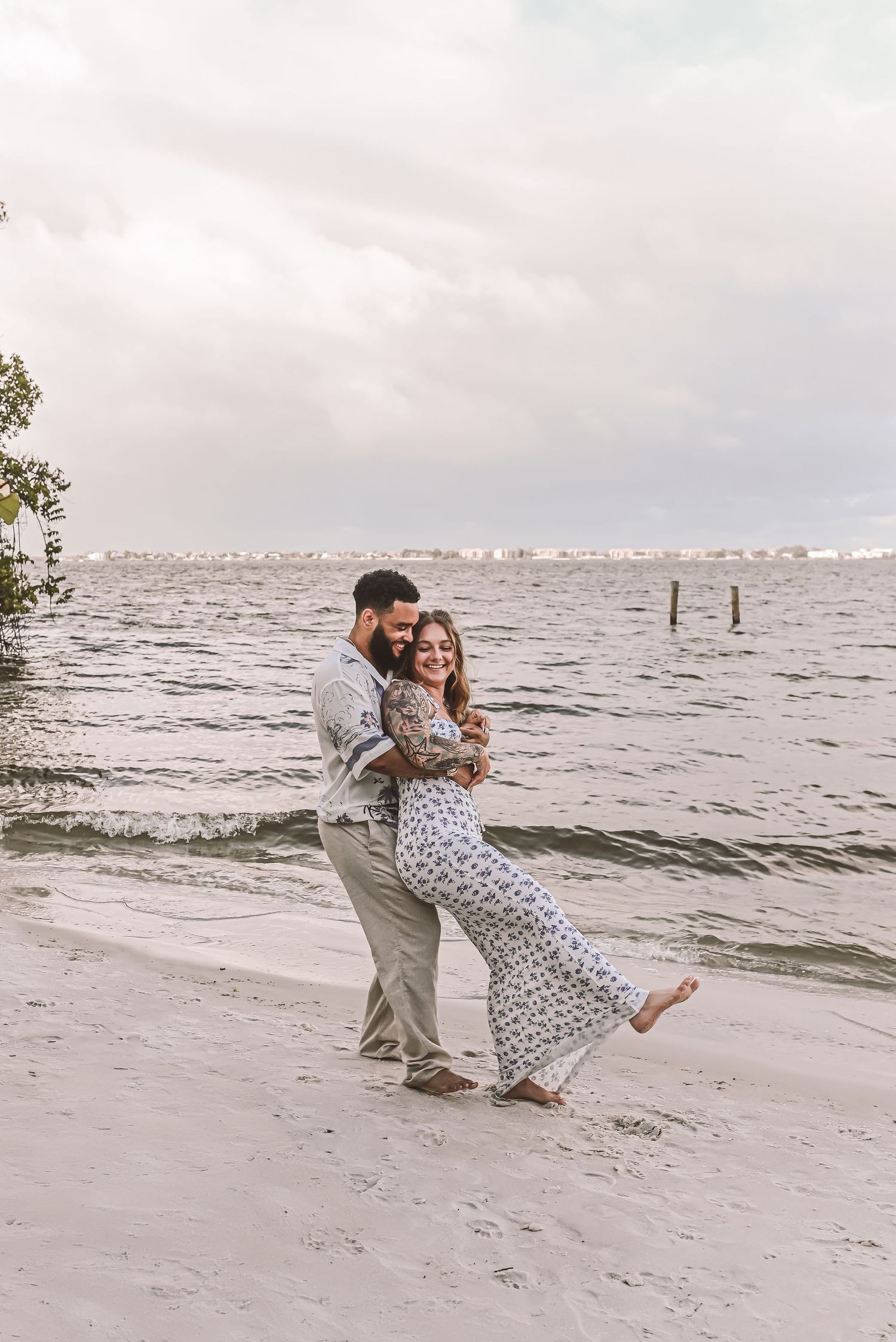 A couple embraces playfully on a cloudy beach shoreline with gentle waves and distant cityscape in the background.