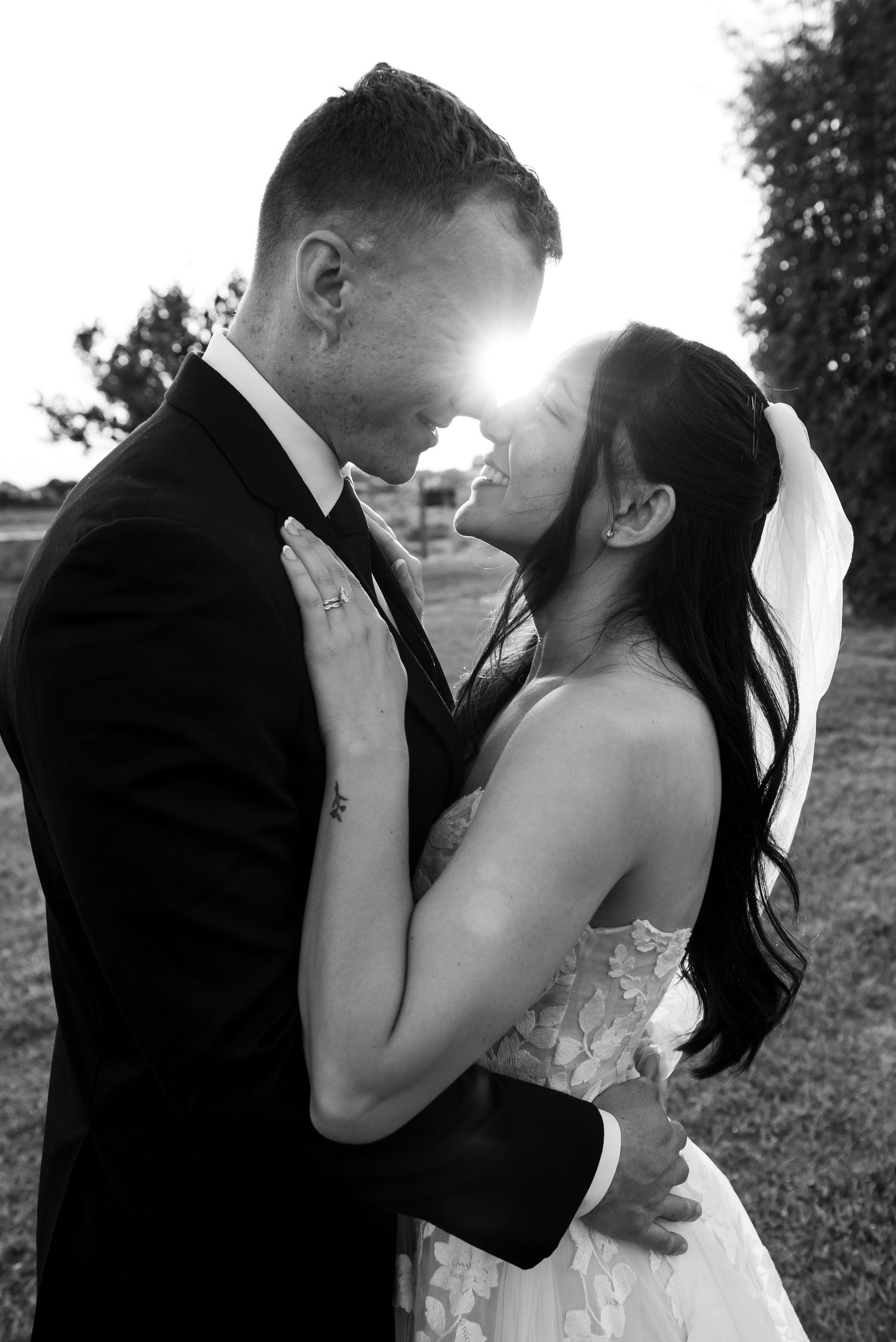 Black and white wedding photo of bride and groom embracing with sunlight flare between them outdoors.