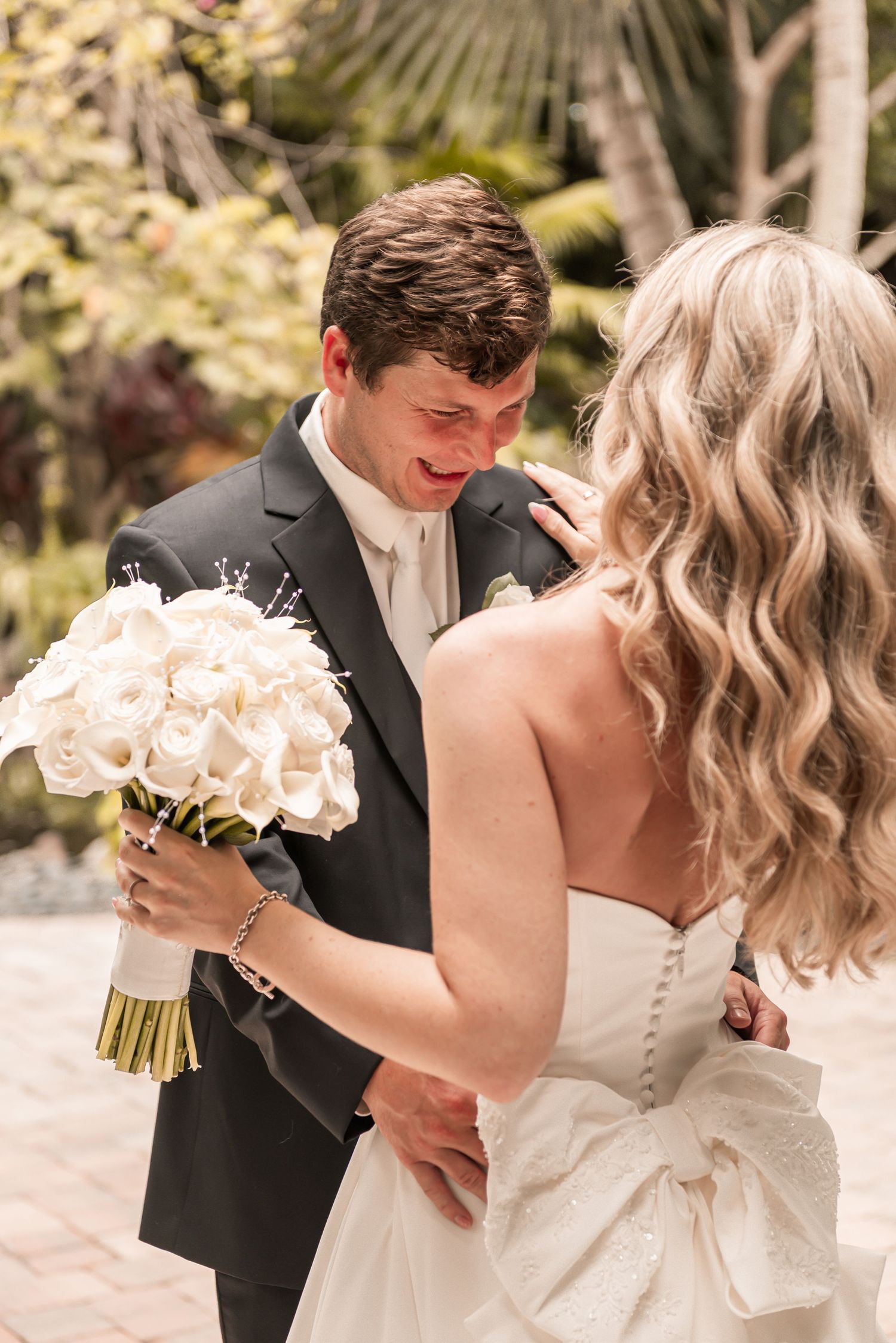 Emotional groom in dark suit tears up seeing bride in white strapless gown holding white rose bouquet outdoors.
