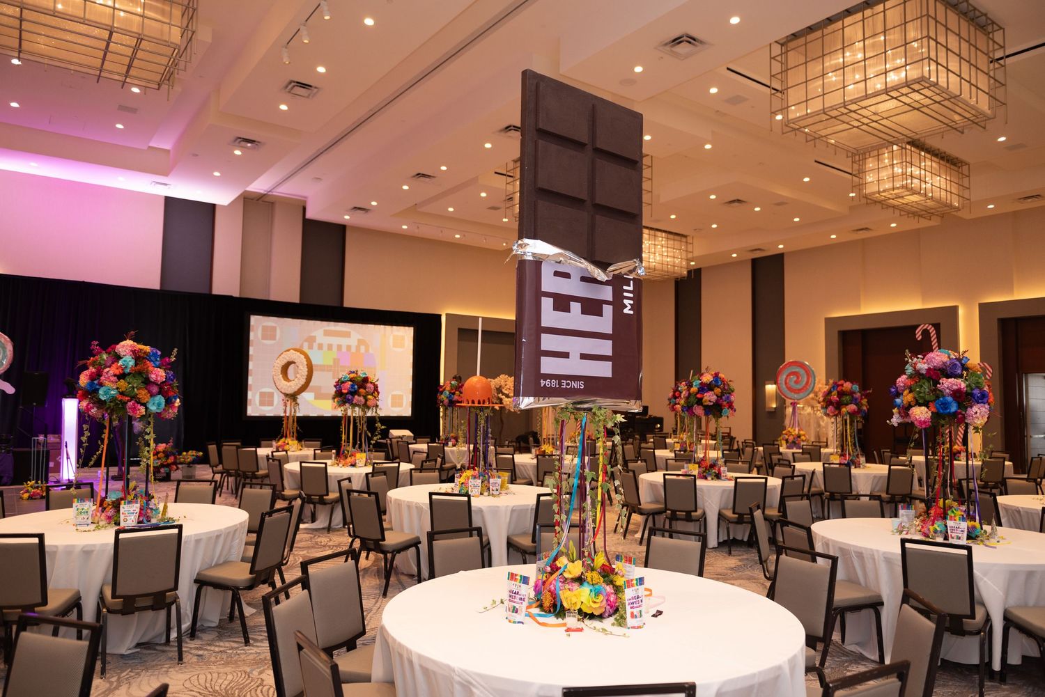 Elegant banquet hall with round tables, floral centerpieces, modern lighting fixtures and stage setup for formal event.