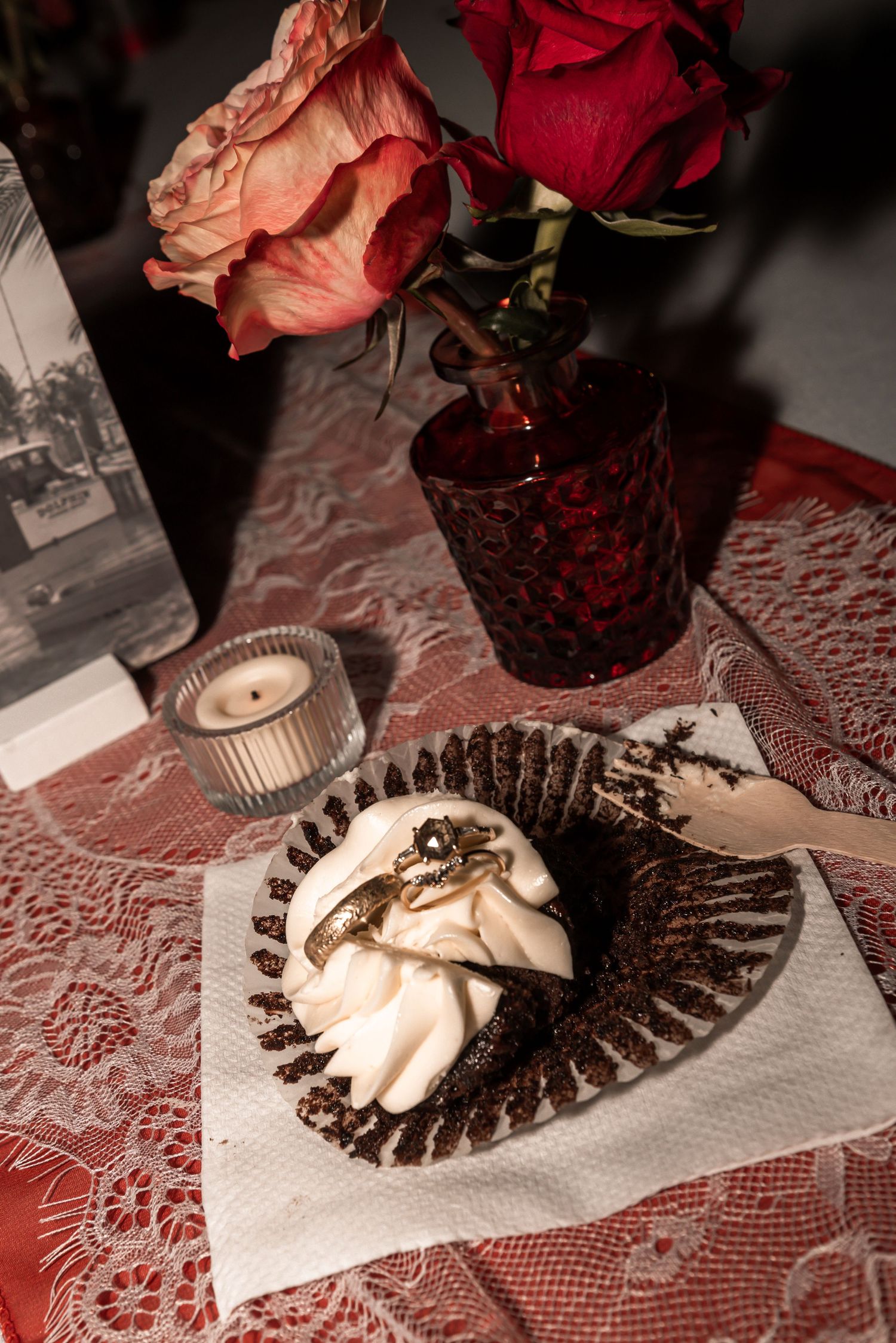 A moody still life with pink and red roses in a dark vase, candle, and decorative dessert on red lace tablecloth.