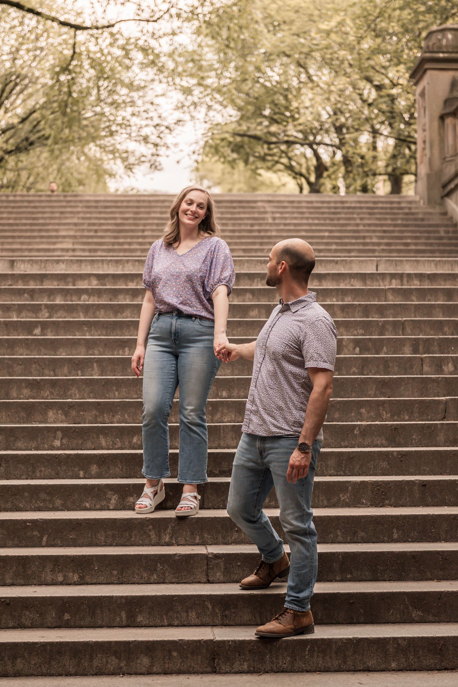 A couple shares a playful moment together while sitting and laughing on outdoor concrete steps surrounded by trees.