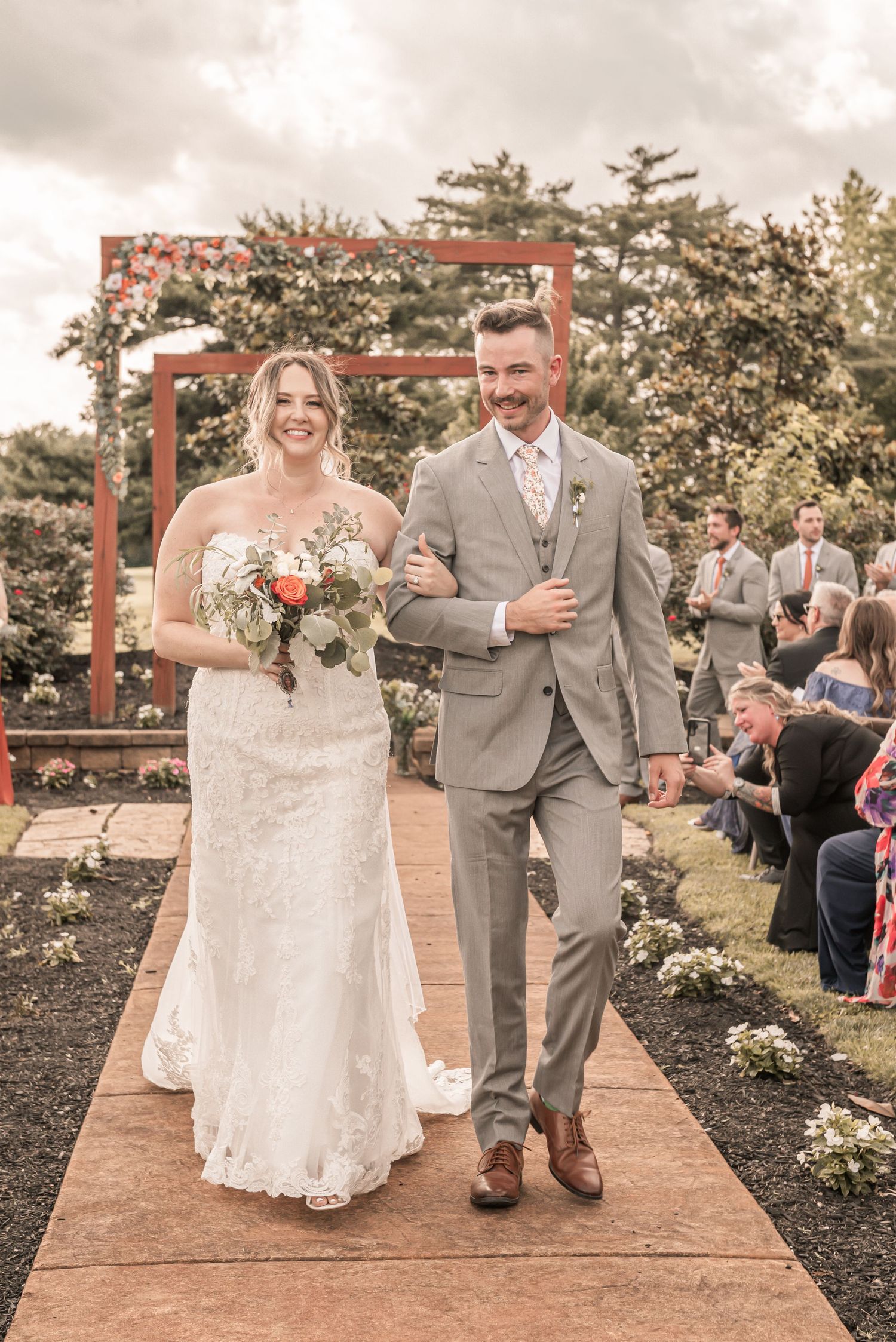 A wedding couple walks down an outdoor aisle under a rustic red arch during a garden ceremony.