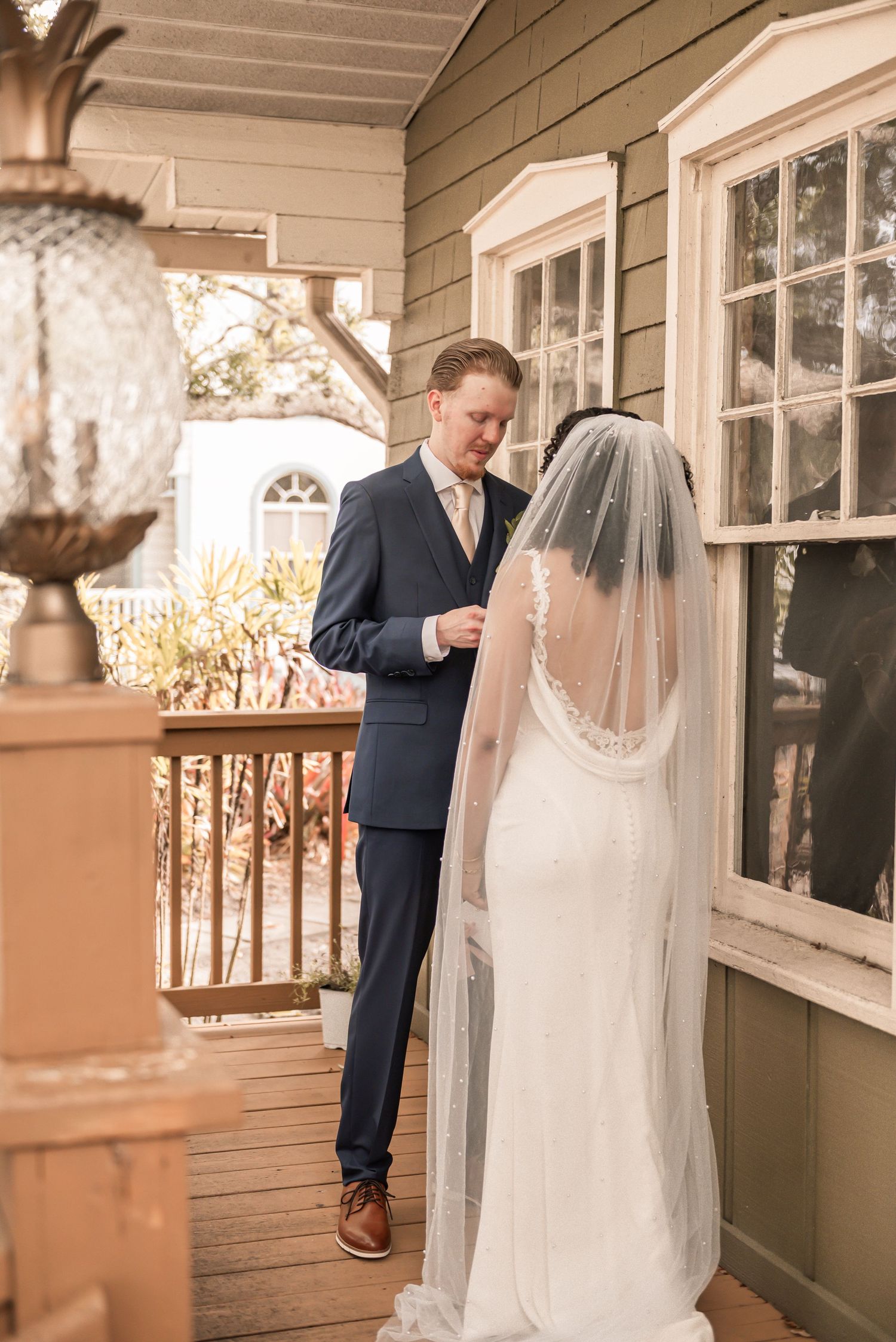 Groom in navy suit reads vows on wooden porch as bride in lace wedding gown and veil stands facing away.