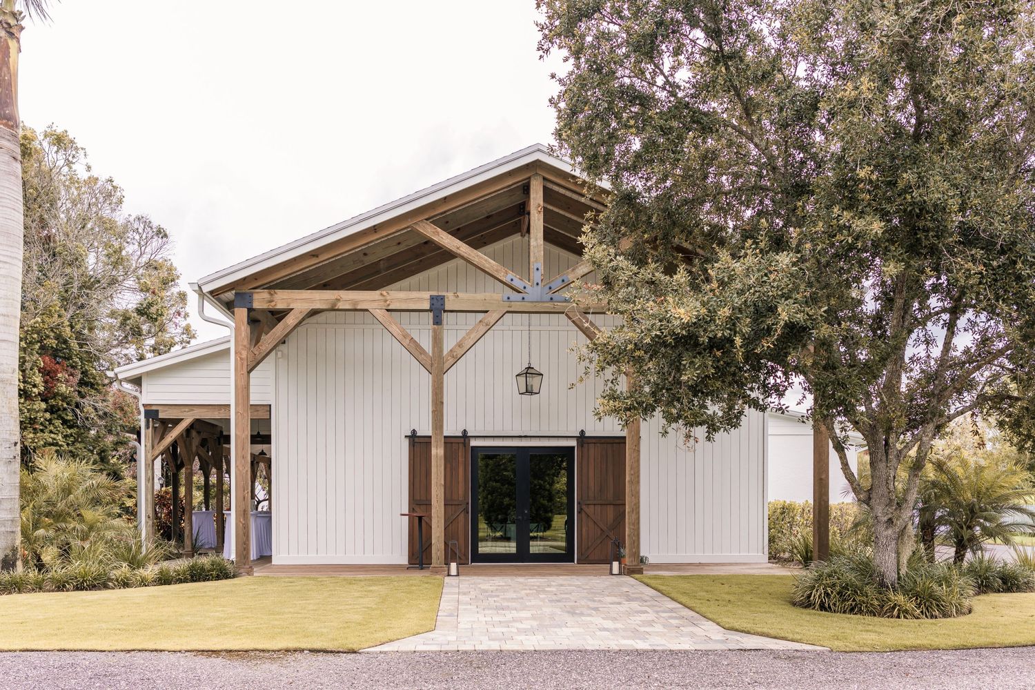 Modern farmhouse barn venue with white board and batten siding, wooden timber frame entrance, and lush oak trees.