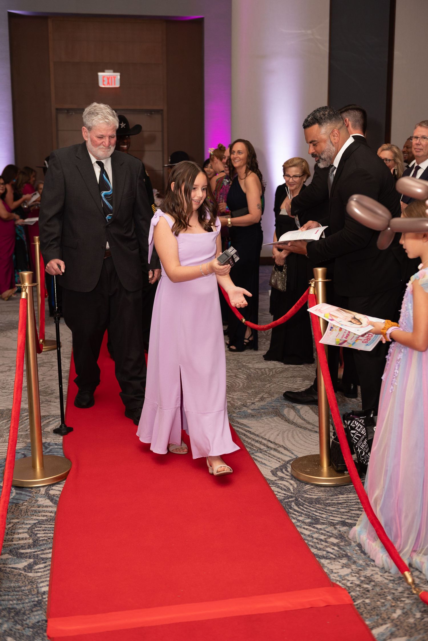 Guests in formal attire walk along a red carpet with velvet ropes at an elegant indoor event venue.