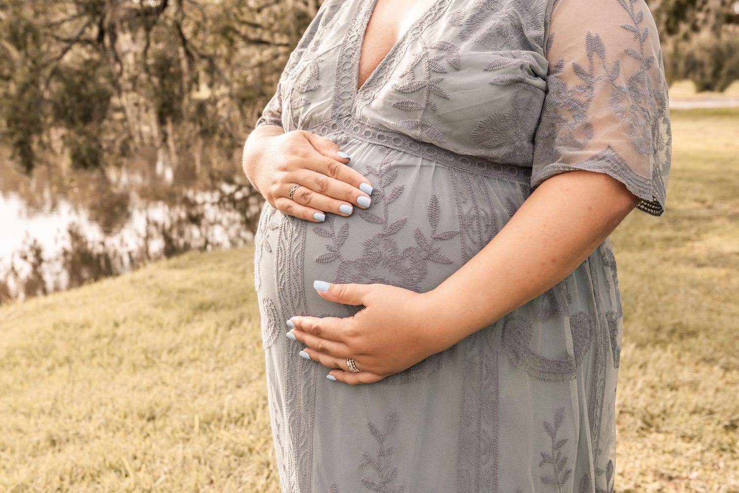 Close up view of pregnant belly in a soft gray floral dress outdoors against a grassy hillside backdrop.