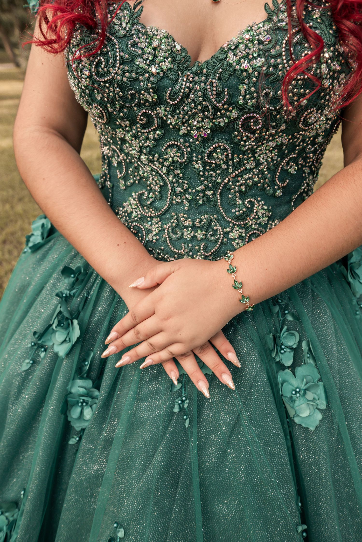 Elegant green quinceañera ball gown with gold beading, 3D floral details, and a sparkling bracelet on folded hands.
