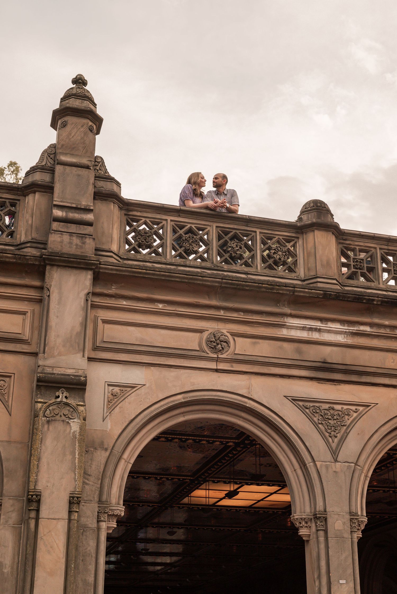 Two figures look down from ornate stone archway with decorative carved details and gothic architecture under cloudy sky.