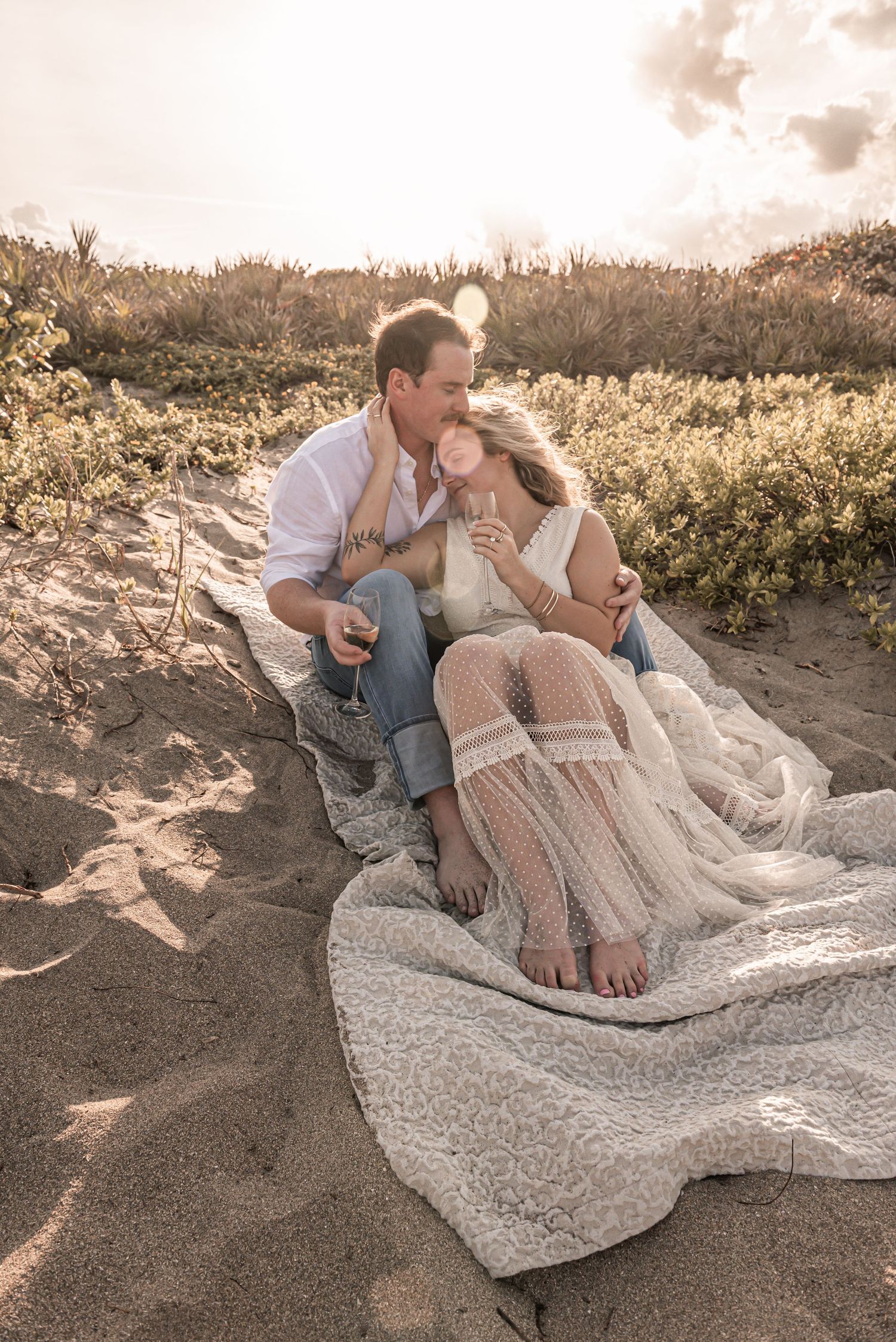 A romantic couple shares an intimate moment on a sandy beach at sunset while resting on a lace blanket.