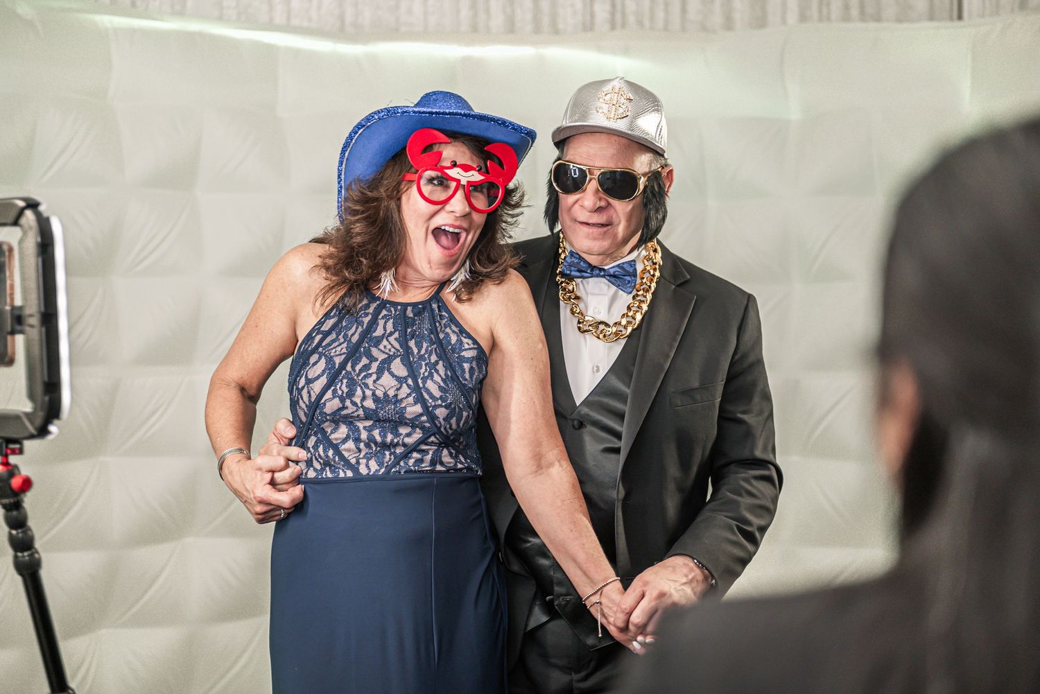 Two party guests have fun posing together in formal attire with a blue hat and sunglasses at a photo booth.