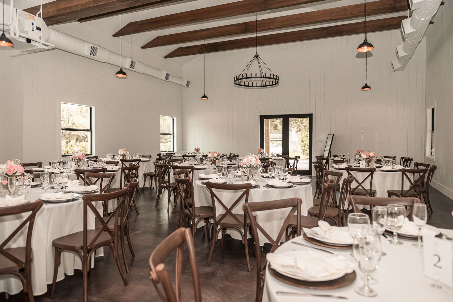 Elegant wedding reception venue with white tablecloths, wooden cross-back chairs, exposed beam ceiling, and chandelier lighting.