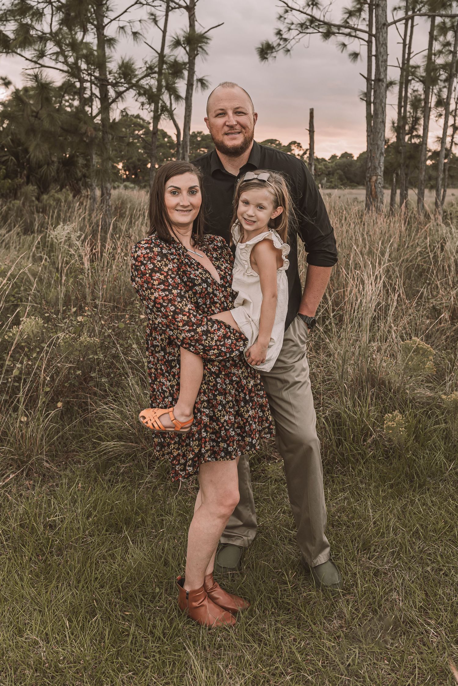 A couple shares playful moments together in a field of tall grass at sunset with pine trees in the background.