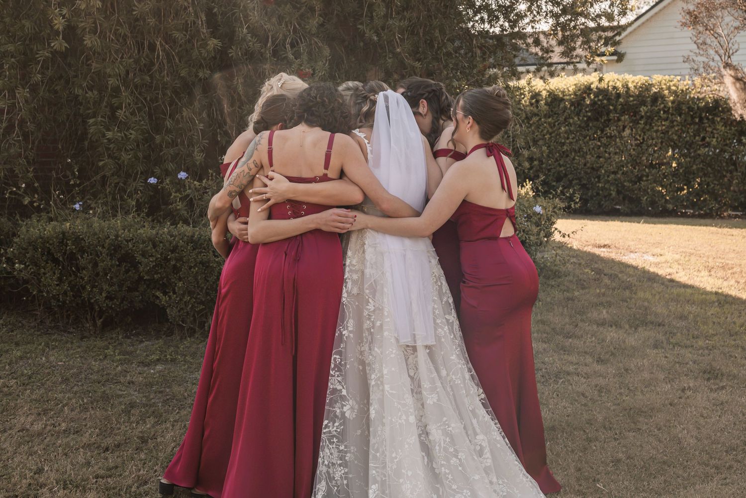 A wedding party in burgundy dresses and white gown pose together during golden hour in an outdoor setting.
